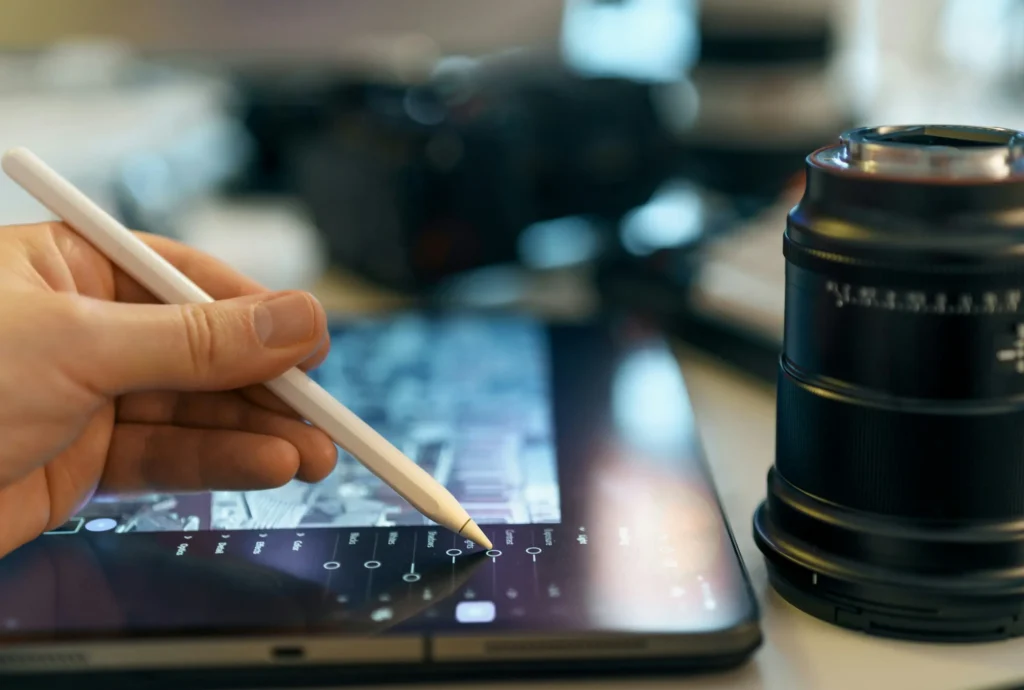 A person using a stylus pen to adjust photo editing sliders (like contrast and light) on a digital tablet next to a camera lens resting on the desk. This image is located in "DIY Photo Editing vs Professional Service" article in Dropicts.com website.
