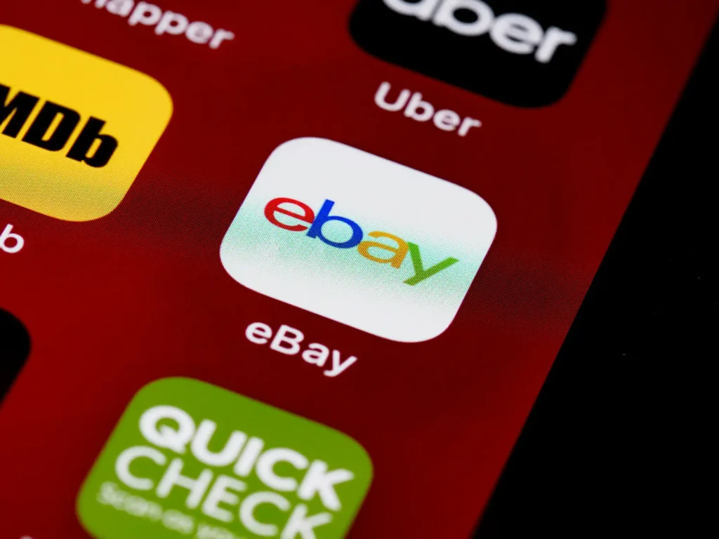 Close-up shot of a smartphone screen displaying the eBay app icon in white with its colorful logo (red, blue, yellow, green) against a red background, surrounded by other app icons like Uber, IMDb, and Quick Check. This is in "Ebay Product Image Requirements" article in Dropicts.com website.