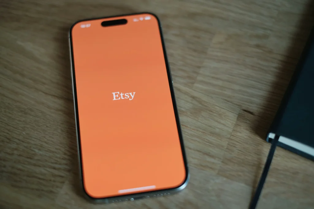 A smartphone lying on a wooden table, displaying the Etsy app with its signature white logo centered on a vibrant orange screen. This image is located in "Etsy Listing Photo Requirements" article in Dropicts.com website.