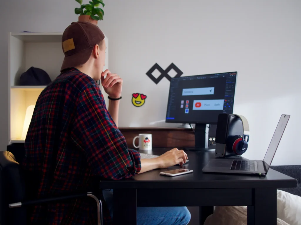 A person wearing a plaid shirt and backward cap sitting at a desk, looking at a computer monitor while working on UI design alongside a laptop and headphones. This images is located in "Freelance Photo Editor vs Photo Editing Company" article in Dropicts.com website.