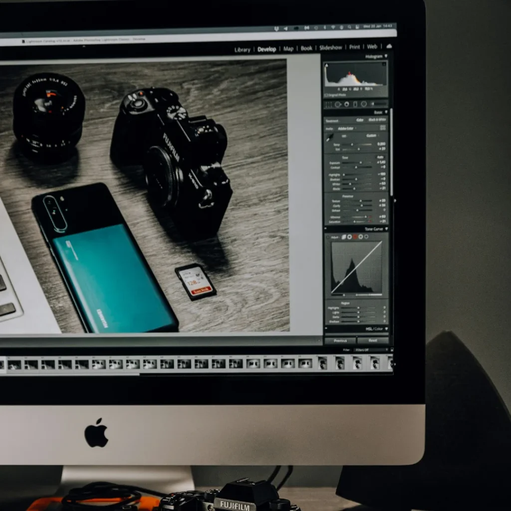 A close-up of an iMac monitor running photo editing software, which displays a picture of a camera, teal smartphone, and SD card on a wooden surface, with a physical Fujifilm camera partially visible resting beneath the screen. This image is located in "Photo Editing Service Free Trial" article in Dropicts.com