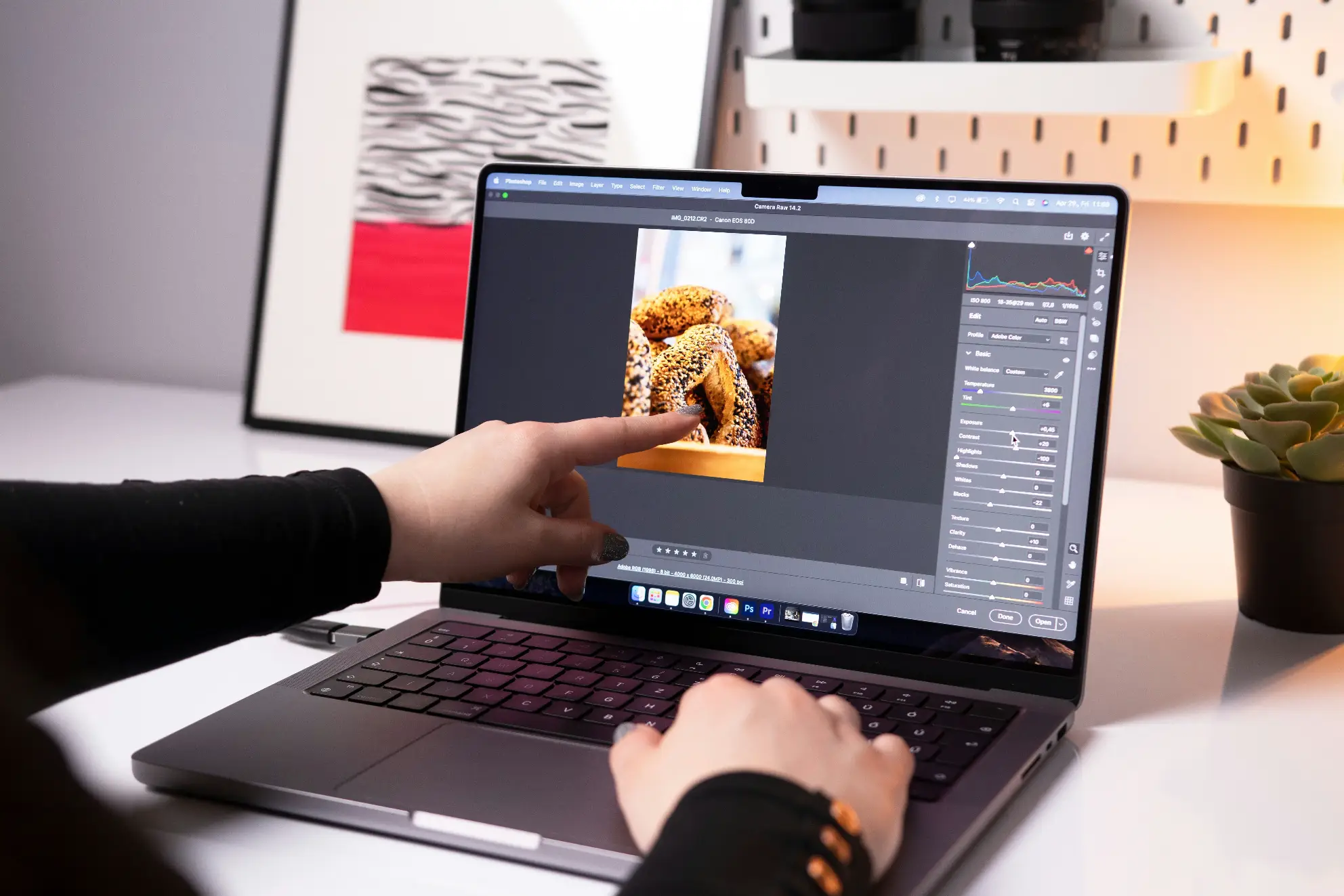 A person editing a food photograph of baked bread rolls on a laptop using Adobe Camera Raw/Photoshop, pointing directly at the image on the screen while adjusting color and exposure sliders on the right panel. This image is located in "Photo Editing Turnaround Time" article in Dropicts.com website.