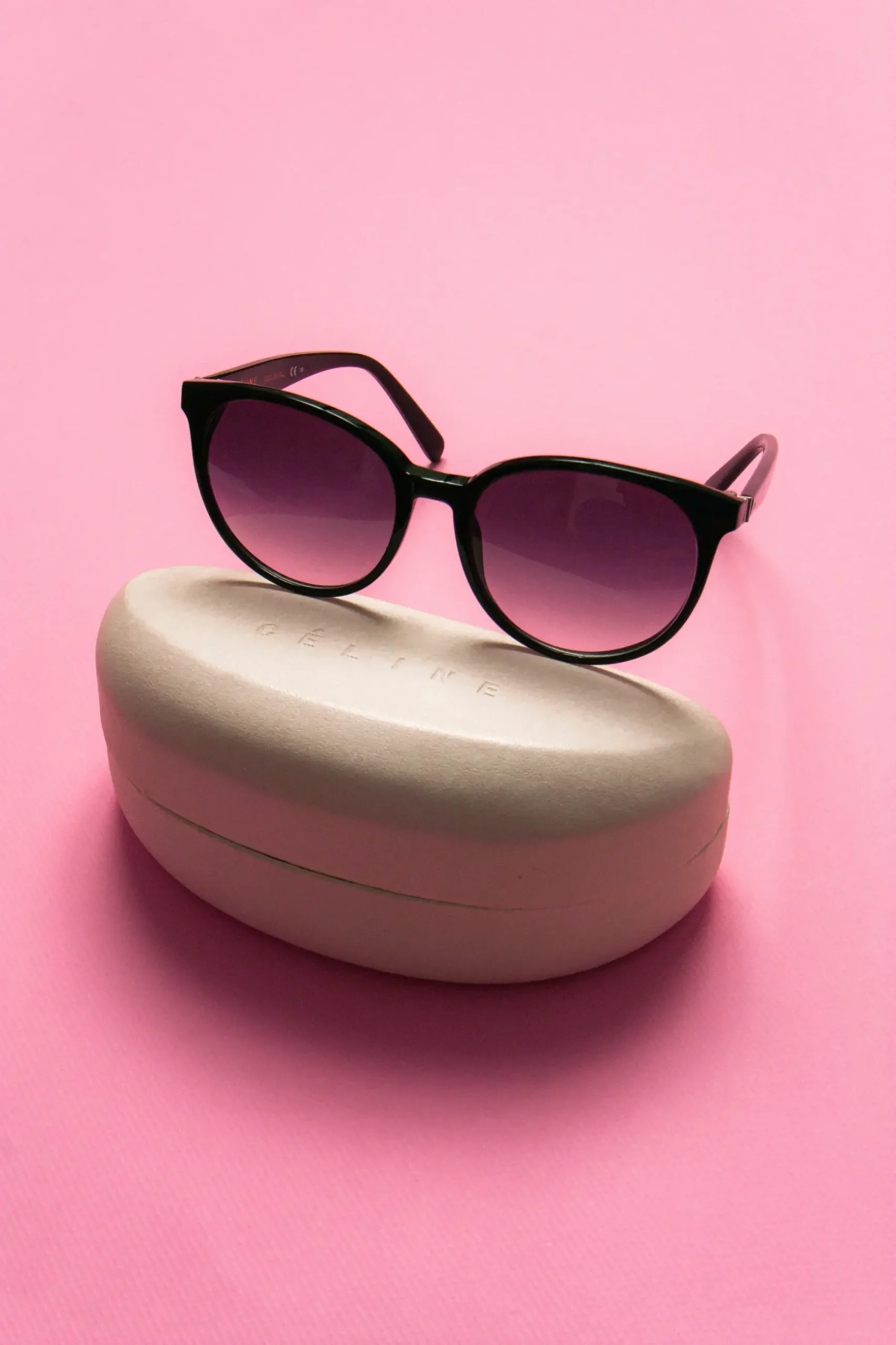 Black sunglasses with purple gradient lenses resting on a light beige Celine-branded hard glasses case, set against a solid pink background. This image is included in "Reverse Image Search Product Photos Scaled" article in Dropicts.com website