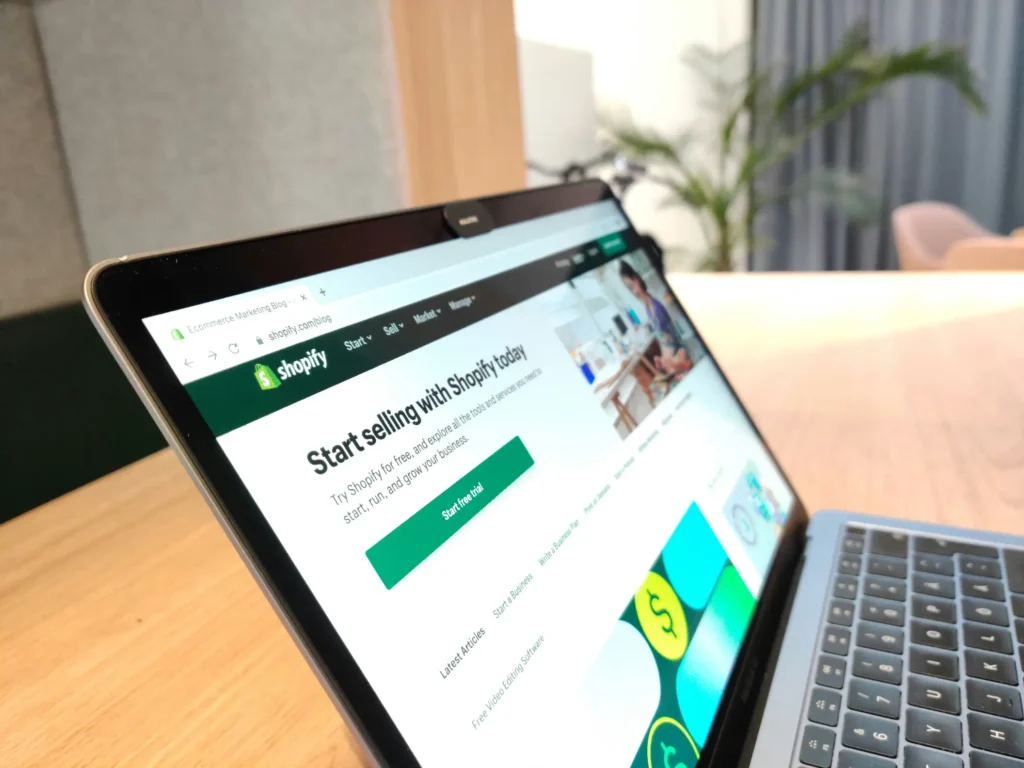 A laptop open on a wooden desk, displaying the Shopify website homepage with the heading "Start selling with Shopify today" and a green "Start free trial" button. This image is located in "Shopify Product Image Size" article in Dropicts.com website.