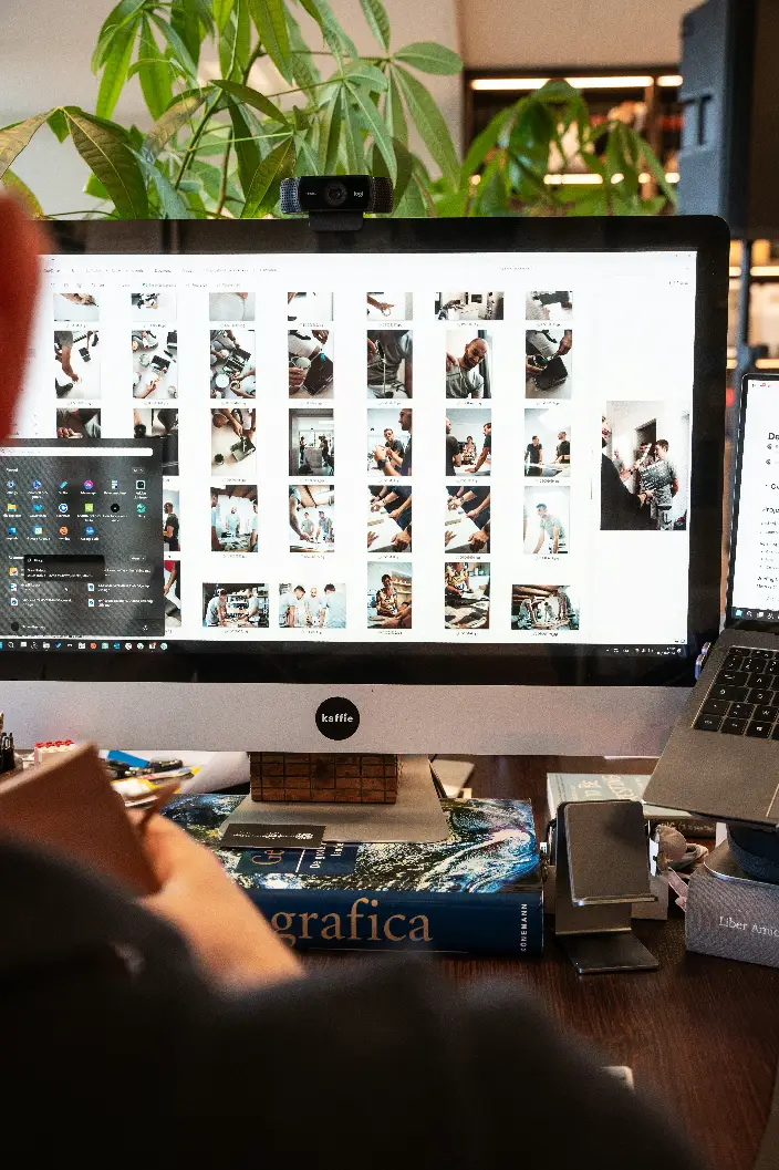 A workspace setup showing a large desktop monitor displaying a photo gallery grid of various photography shots, with a webcam mounted on top, surrounded by books and office items on the desk. This image is located in "Switch Photo Editing Service" article in Dropicts.com website.