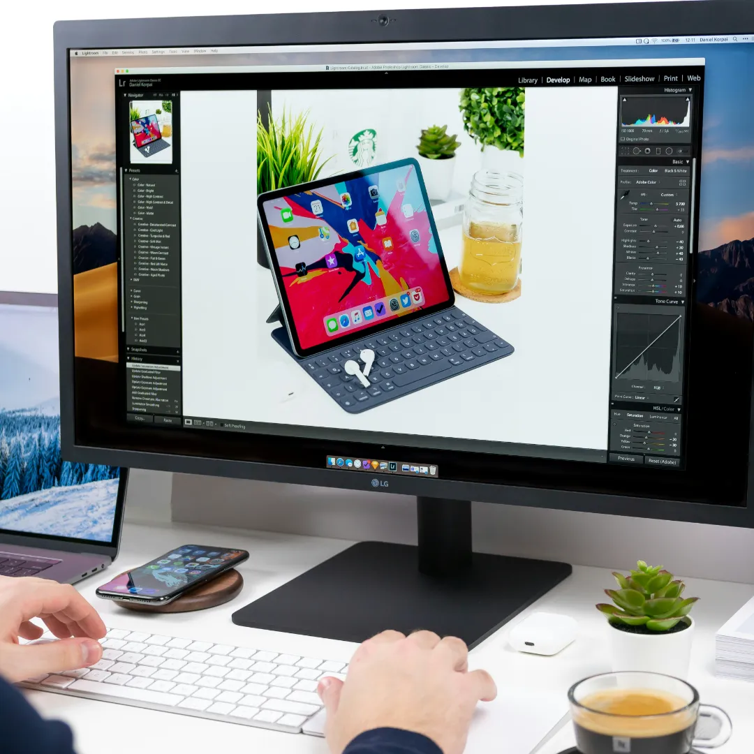 A person's hands using a white keyboard at a bright desk setup in front of an LG monitor; the screen displays Adobe Lightroom showing an edited product photo of an iPad Pro, keyboard case, and AirPods. This image is located in "Unlimited Revisions Photo Editing Service" article in Dropicts.com website.