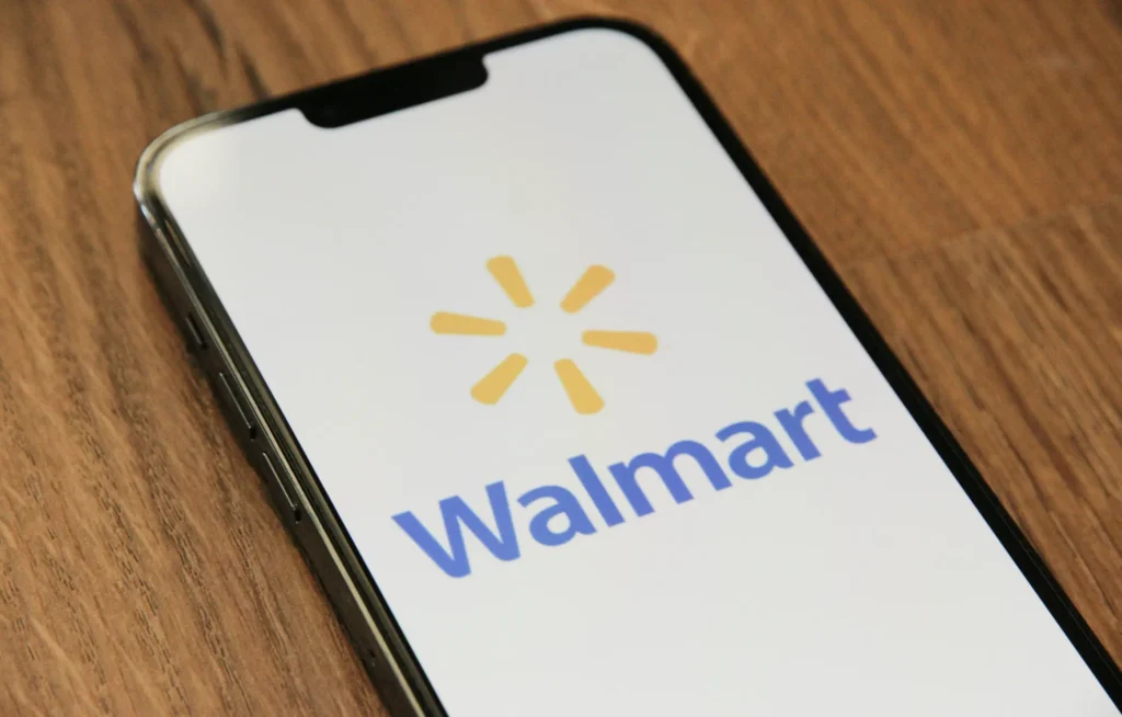 A smartphone lying flat on a wooden surface, displaying the Walmart logo and text in blue and yellow on its screen. This is in "Walmart Marketplace Product Image Requirements" article in Dropicts.com website.